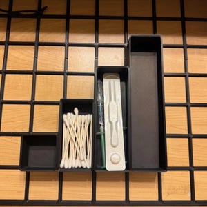 May include: A black organizer with four compartments, containing cotton swabs, a syringe, and a medical device. The organizer is set against a grid pattern with a wooden background.