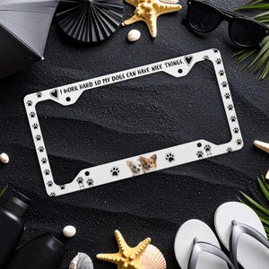 May include: White license plate frame with black paw prints and the text "I WORK HARD SO MY DOGS CAN HAVE NICE THINGS." Features two small dogs and a heart. A decorative accessory for dog lovers.