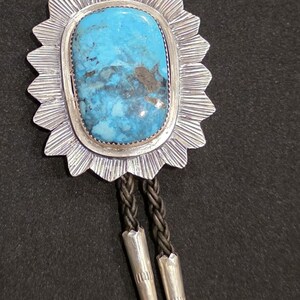 May include: A silver bolo tie with a turquoise stone set in a starburst design. The bolo tie has a black braided cord.
