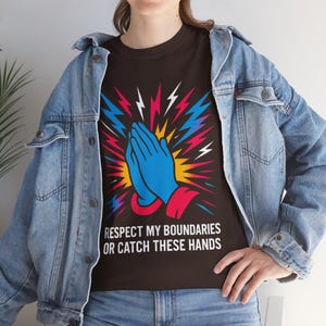 May include: Black t-shirt featuring a graphic of two blue hands in a praying position with a yellow, red and blue lightning bolt design. The text "Respect my boundaries or catch these hands" is printed below the graphic.
