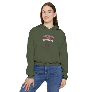 Könnte beinhalten: Ein olivgrüner Kapuzenpullover mit dem Text "Clocked In and Clueless" in Rosa. Der Pullover hat einen Kordelzug in der Taille und lange Ärmel. Die Grafik auf dem Sweatshirt ist eine Strichzeichnung einer Person, die einen Klemmbrett hält.