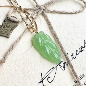 Puede incluir: Collar de cadena dorada con un colgante en forma de hoja verde claro. El colgante presenta detalles de vetas. Se muestra sobre un fondo de papel texturizado con un pequeño dije en forma de corazón y cuerda.