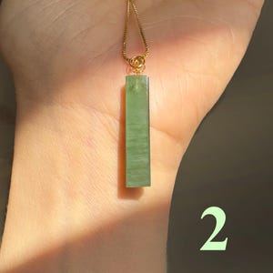 Puede incluir: Un colgante rectangular de jade verde claro cuelga de una delicada cadena dorada. El jade tiene una superficie lisa y pulida y una sutil variación de color natural. El colgante está suspendido de un pequeño y ornamentado cierre dorado.