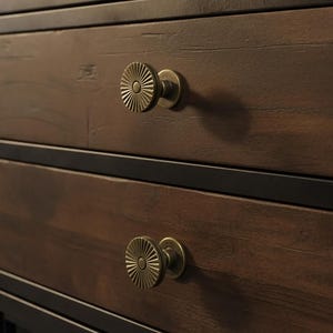 Puede incluir: Primer plano de una vinoteca de madera con puertas de cristal y molduras metálicas. El mueble tiene cajones con tiradores decorativos de latón. Se ven botellas de vino dentro del mueble y en una balda superior.