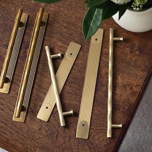 Puede incluir: Una colección de herrajes de latón para armarios, que incluye tiradores largos y tiradores en T, dispuestos sobre una superficie de madera oscura. Los tiradores tienen una variedad de diseños, incluyendo superficies acanaladas y texturizadas. Un pequeño jarrón blanco con vegetación en el fondo.