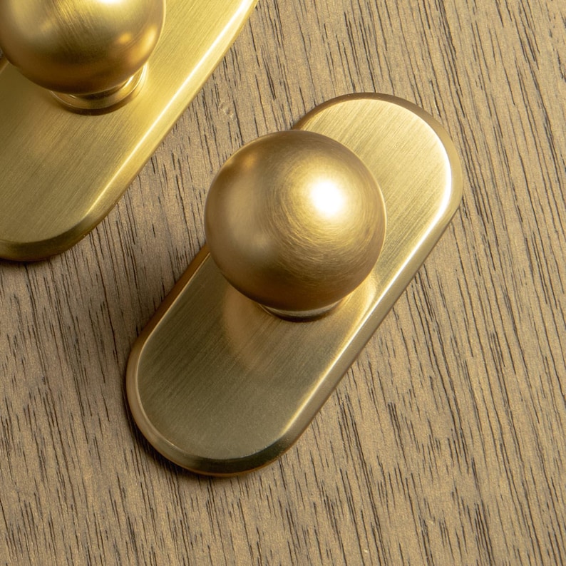 May include: Close-up of two brass cabinet knobs. Each knob features a rounded, spherical handle atop a rectangular backplate with rounded edges. The knobs have a brushed finish and are set against a wood grain surface.
