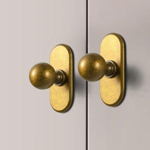 May include: Two antique brass cabinet knobs. Each knob features a spherical handle attached to an elongated, oval backplate. The hardware has a warm, aged finish, suggesting a vintage aesthetic. The knobs are mounted on a light gray surface.