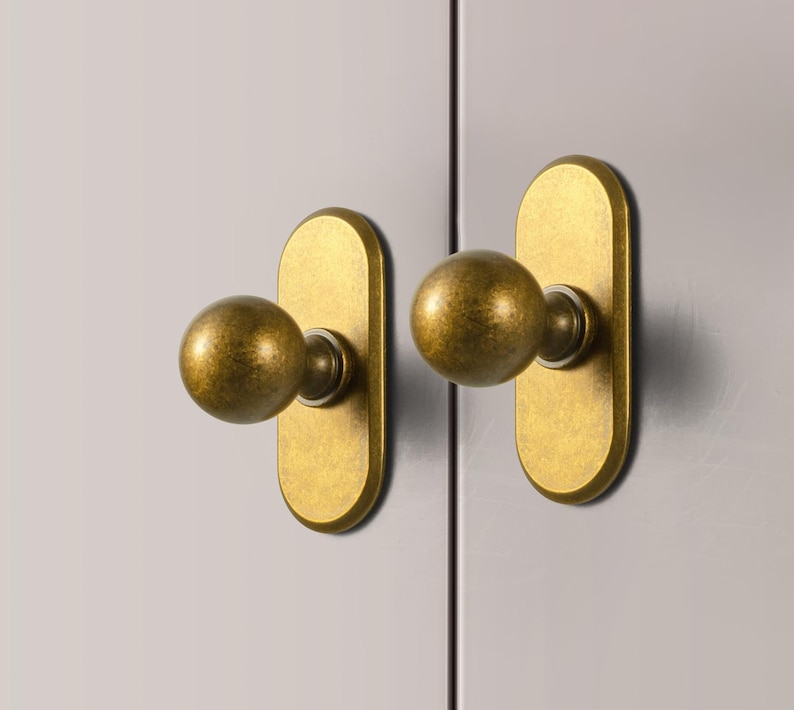 May include: Two antique brass cabinet knobs. Each knob features a spherical handle attached to an elongated, oval backplate. The hardware has a warm, aged finish, suggesting a vintage aesthetic. The knobs are mounted on a light gray surface.