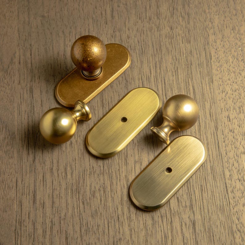 May include: Four brass cabinet knobs and pulls on a wooden surface. Two knobs have a spherical design, and two have an oval backplate. The hardware has a warm, golden finish. The wood grain is visible in the background.