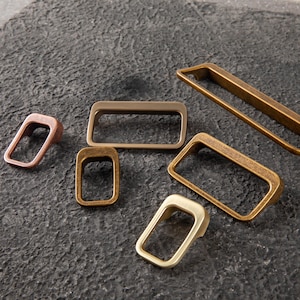 May include: Various rectangular cabinet pulls in bronze, copper, and brass tones are arranged on a dark, textured surface. The pulls vary in size and shape, with some having a handle design.