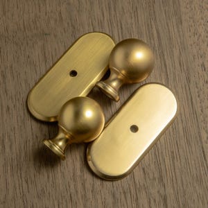 May include: Four brass-colored furniture hardware pieces. Two are oval-shaped with a single hole, and two are spherical finials with a decorative base. The hardware has a brushed finish and is set against a wood grain background.