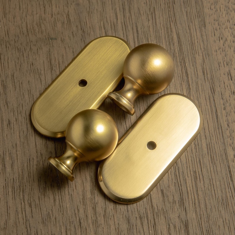 May include: Four brass-colored furniture hardware pieces. Two are oval-shaped with a single hole, and two are spherical finials with a decorative base. The hardware has a brushed finish and is set against a wood grain background.