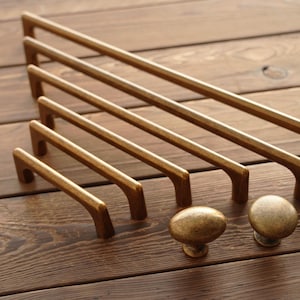 May include: A collection of antique brass cabinet hardware, including several sizes of bar pulls and two round knobs. The hardware is arranged on a wooden surface, showcasing the warm, aged finish and design.