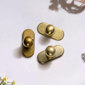 May include: Three brass cabinet knobs with a gold finish. Each knob features a rounded, spherical handle attached to a rectangular backplate. The knobs are arranged on a white surface, with soft shadows and a few flowers in the corner.