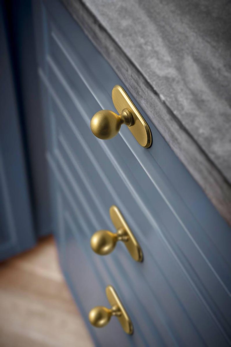 May include: Close-up of a blue cabinet with three gold-colored knobs. The knobs have a rounded shape and are mounted on rectangular backplates. The cabinet has a grooved design.