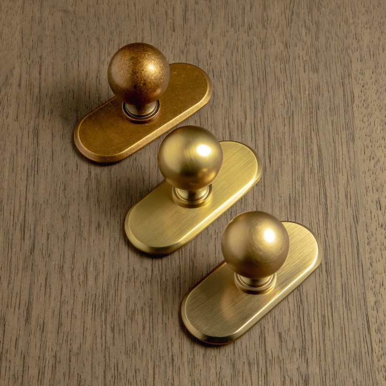 May include: Three brass cabinet knobs with a ball-shaped handle and an oval backplate. The knobs are in varying shades of gold, from a darker, antique finish to a bright, polished brass. The knobs are arranged on a wooden surface.