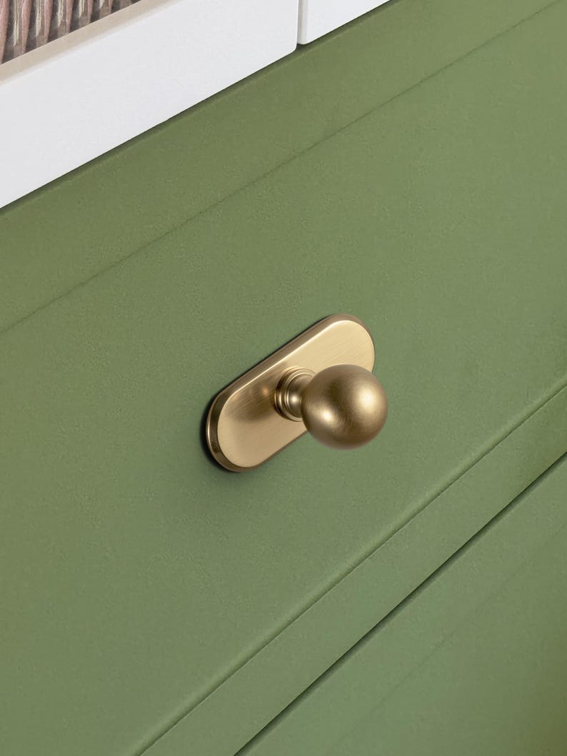 May include: Close-up of a gold-toned cabinet knob with a rounded handle and a rectangular base. The knob is mounted on a green cabinet door. The cabinet door has a recessed panel design.