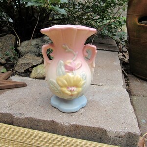 May include: A small, pink and blue ceramic vase with floral designs. The vase has two handles and a scalloped rim.