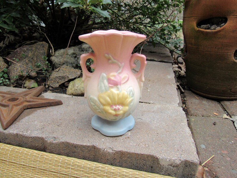 May include: A small, pink and blue ceramic vase with floral designs. The vase has two handles and a scalloped rim.