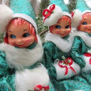May include: Three vintage Christmas elf dolls with green sparkly dresses, white fur trim, and red ribbon accents. The elves have white fur hats with red and white accents.