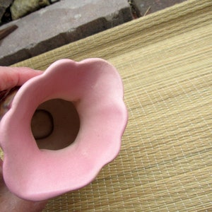 May include: A pink ceramic vase with a wide opening and a scalloped rim. The vase is sitting on a woven mat.