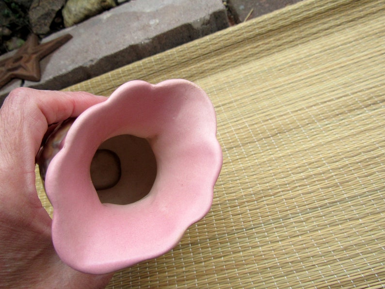 May include: A pink ceramic vase with a wide opening and a scalloped rim. The vase is sitting on a woven mat.