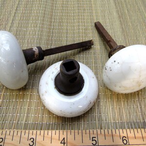 May include: Three antique white porcelain door knobs with aged, rusty metal hardware. The knobs are oval-shaped and show signs of wear, with a square base and a ruler in the foreground.