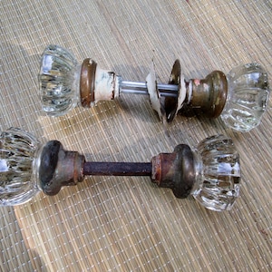 May include: Two sets of antique glass doorknobs with brass hardware. One set has a rusty metal rod connecting the knobs, while the other set has a separate rod and a white metal plate.