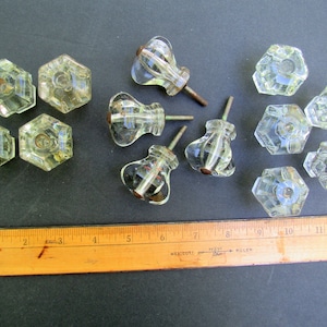 May include: A collection of 10 clear glass cabinet knobs, including 6 hexagonal knobs and 4 round knobs with a fluted design. The knobs have a silver metal screw and are arranged on a dark surface with a wooden ruler for scale.