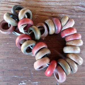 May include: A necklace of wooden rings in various colors, including red, green, and natural wood tones. The rings are strung together on a white cord, creating a circular shape. The background is a weathered wooden surface.