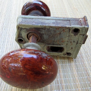 May include: A vintage door knob with a brown ceramic handle and a metal lock plate. The lock plate is rusty and has a keyhole.