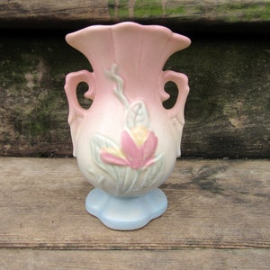 May include: A small, pink and blue ceramic vase with a floral design. The vase has two handles and a scalloped rim.