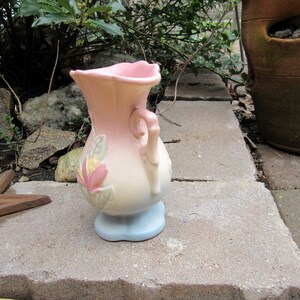 May include: A small, pink and white ceramic vase with a floral design and a handle. The vase has a blue base.
