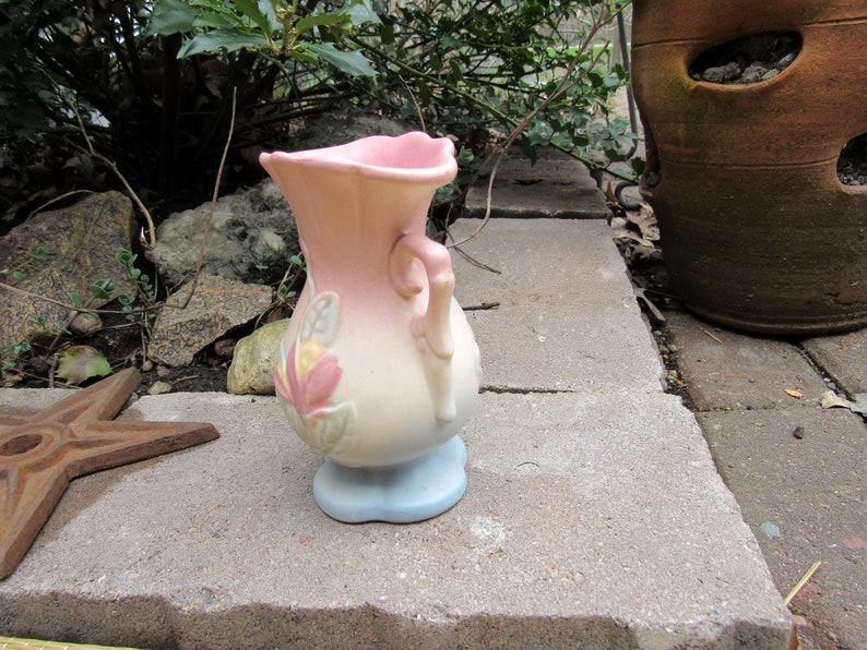 May include: A small, pink and white ceramic vase with a floral design and a handle. The vase has a blue base.