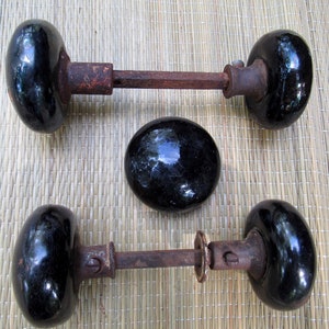 May include: Three antique black door knobs with a glossy finish and aged, rusty metal hardware. The knobs are oval-shaped, and one is a single sphere. The door knobs are likely vintage or reclaimed.