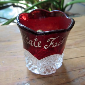 May include: A small, clear glass cup with a red glass top. The top has a scalloped edge and the words "State Fair" are written in white on the red glass.