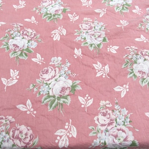 May include: A dusty rose fabric with a repeating floral pattern. Clusters of white and pink roses, accented with green leaves and small blue flowers, are scattered across the surface. White leaf sprigs fill the spaces.