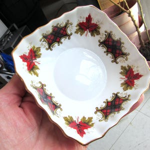 May include: A white square ceramic dish with a gold rim and a red, black, and green plaid pattern. The dish is decorated with red maple leaves and gold accents.