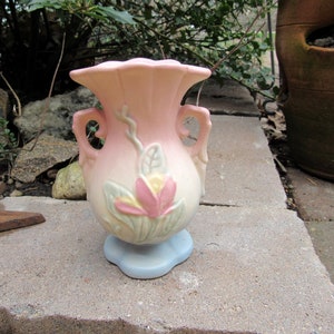 May include: A small, pink and white ceramic vase with a floral design. The vase has two handles and a scalloped rim.