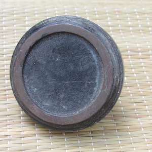 May include: A dark, round object with a textured, black center and a brown, circular rim. The object appears to be made of a dark material, possibly metal or ceramic. The object is sitting on a woven, light-colored surface.