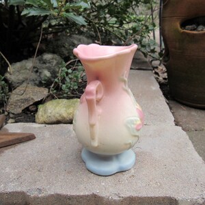 May include: A pink and white ceramic vase with a floral design and a handle. The vase has a light blue base.
