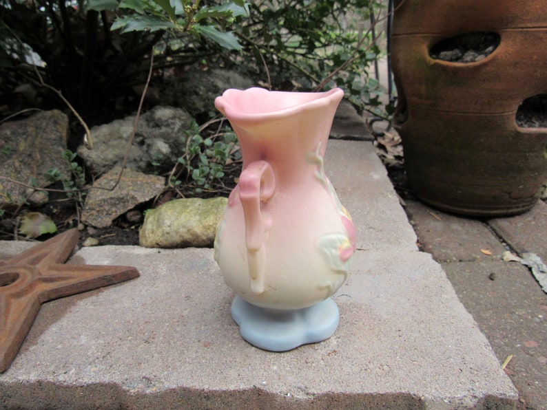 May include: A pink and white ceramic vase with a floral design and a handle. The vase has a light blue base.