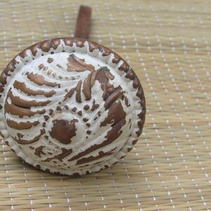May include: A round, antique-style bottle stopper with a decorative design. The stopper features a white and brown color scheme with a detailed relief pattern and a small handle. The design includes a horse-like figure.