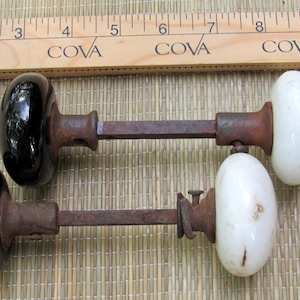 May include: Two antique door knobs with oval-shaped porcelain handles. One is black, the other is white. The knobs are connected by a rusty metal rod. A wooden ruler with the word "COVA" is in the background.