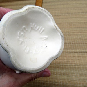 May include: The bottom of a white ceramic vase with a scalloped edge. The vase has the words "Plastic Hull" printed on the bottom.