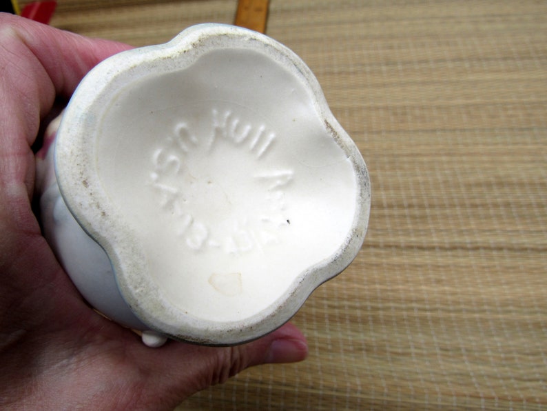 May include: The bottom of a white ceramic vase with a scalloped edge. The vase has the words "Plastic Hull" printed on the bottom.