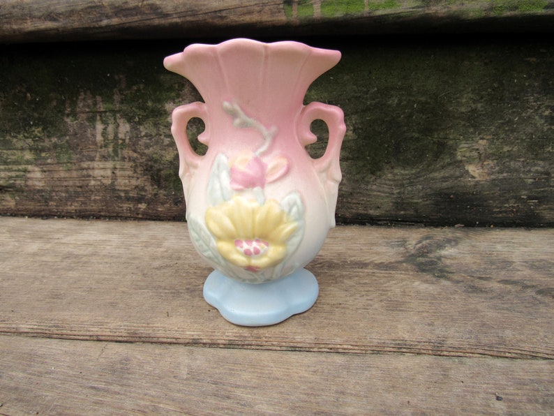 May include: A small, pink and blue ceramic vase with floral designs. The vase has two handles and a scalloped base.