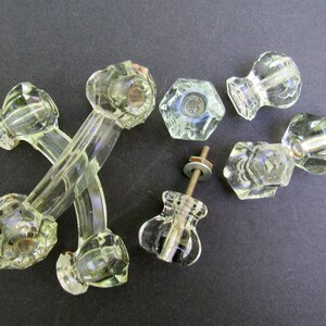 May include: A set of eight clear glass cabinet knobs, including two pairs of ornate handles with a cross-shaped design, two round knobs, and two hexagonal knobs. The knobs are in various styles and sizes, with some featuring intricate details.