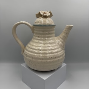 May include: Cream-colored ceramic teapot with a flower-shaped lid handle. The teapot has a curved handle and spout, with a textured, speckled surface. The lid has a light blue rim. The teapot is displayed on a white square pedestal.