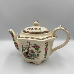 May include: Cream-colored ceramic teapot with a floral design. The teapot features a lid with a small knob and a curved handle. The floral pattern includes pink and red flowers with green leaves.
